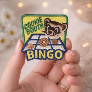 May include: Embroidered patch with a yellow background and green border. The patch features a cartoon ferret with the words "COOKIE BOOTH" and "BINGO". The ferret is brown and white, and the bingo board has brown cookies.