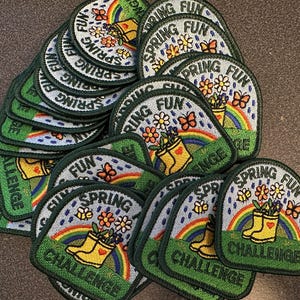 May include: A collection of embroidered patches with a spring theme. Each patch features a rainbow, yellow rain boots, flowers, and butterflies. The text "SPRING FUN CHALLENGE" is included on each patch, with a green border.