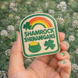 May include: A St. Patrick's Day patch with a rainbow, shamrocks, and a pot of gold. The patch reads "Shamrock Shenanigans" in green text. The patch has a green border and is held in a hand.