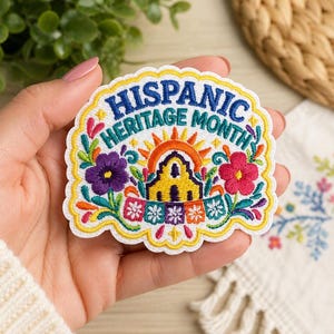 May include: An embroidered patch with a scalloped edge featuring the text "HISPANIC HERITAGE MONTH" in blue. The design includes a yellow building, an orange sun, a purple flower, a pink flower, and colorful papel picado banners on a white background.