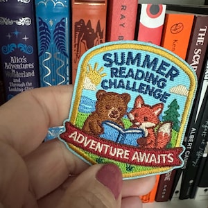 May include: Embroidered patch with a light blue background featuring a bear and fox reading a book. The patch reads "Summer Reading Challenge" and "Adventure Awaits" in white and red lettering. A sun and trees are also depicted.