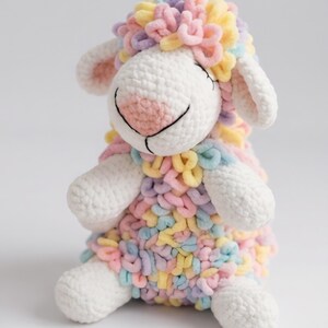 May include: A plush stuffed animal shaped like a sheep with a white face, ears, arms, and feet. The body and head are covered in textured, looped yarn in pastel shades of pink, yellow, blue, and lavender. It has a stitched pink nose and eyes.