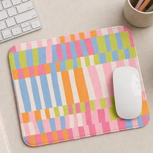May include: A rectangular mousepad with a colorful abstract design. The pattern features vertical stripes in shades of pink, blue, orange, and green. A white computer mouse rests on the pad, next to a keyboard and a pen.