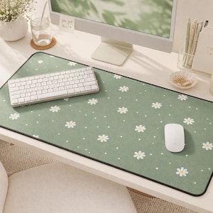 May include: A green desk mat with a daisy pattern, a white keyboard, and a white mouse. The mat has a black border and is on a white desk. A computer monitor, a candle, and stationery are also visible.