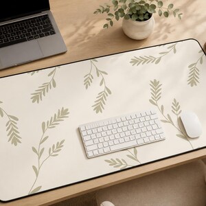 May include: A cream-colored desk mat with a black border, featuring a repeating pattern of olive-green botanical sprigs. A laptop, keyboard, and mouse are on the mat, along with a cup of coffee and a small plant.