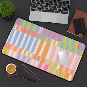May include: A rectangular desk mat with a colorful geometric pattern. The design features vertical and horizontal stripes in shades of pink, orange, blue, and green. A white computer mouse sits on the mat, with a laptop and notepad in the background.