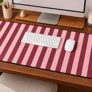 May include: A desk mat featuring a striped design in alternating pink and burgundy shades. A white keyboard and mouse are placed on the mat. The mat sits on a wooden desk, with a computer monitor, a candle, and a coffee cup in the background.