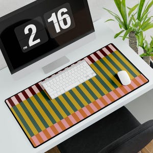 May include: A desk mat with a striped design in green, yellow, pink, and burgundy. A white keyboard and mouse are on the mat, with a computer monitor behind. The monitor displays the time as 2:16 PM. The mat is designed for a desk.