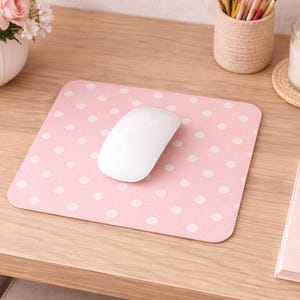 May include: A pink mousepad with white polka dots and a white wireless mouse sits on a wooden desk. A white candle in a glass holder, a pink notebook, and a gold pen are also on the desk. A vase of pink roses is in the background.