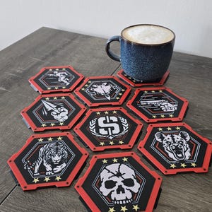 May include: A set of hexagonal coasters featuring diverse graphic designs, such as a tiger and a skull, with red and black borders. A blue mug containing a coffee drink rests on one of the coasters. The coasters are displayed on a wooden surface.