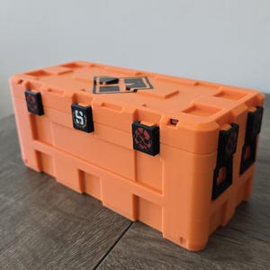 Könnte beinhalten: Ein orangefarbener rechteckiger Aufbewahrungsbehälter mit schwarzen Akzenten. Der Behälter hat eine Reihe schwarzer Verschlüsse und ein Logo auf der Oberseite. Die Verschlüsse haben Schädel- und Buchstabenmuster. Der Behälter besteht aus einem robusten Material.