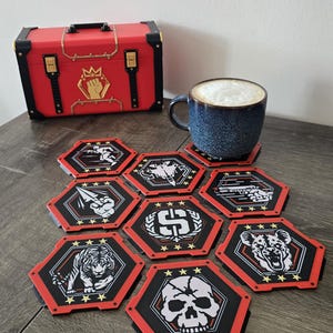 May include: A set of seven hexagonal coasters featuring black, red, and white graphic designs of a skull, tiger, hyena, jet, fist, and currency symbol, arranged around a blue ceramic mug filled with a foamy beverage near a red storage chest.