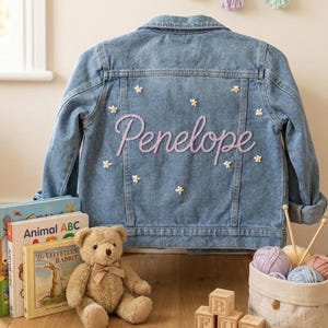 Puede incluir: Chaqueta vaquera azul claro con el nombre "Penelope" bordado en cursiva en la espalda. La chaqueta está decorada con pequeñas aplicaciones de flores blancas. Un osito de peluche, libros y bloques de madera están en primer plano.