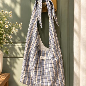 May include: A blue and white plaid hobo bag with a knotted shoulder strap. The bag features a white fabric label with the text saki studio printed in black. The interior lining is a solid white fabric, and the exterior has a grid pattern.