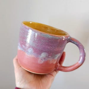 Puede incluir: Una taza de cerámica hecha a mano con un degradado de color único. La taza pasa de una base rosa coral a una sección media lavanda y azul claro, culminando en un interior amarillo dorado. Tiene un asa redondeada.