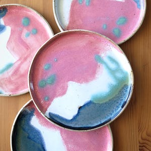 Może przedstawiać: Cztery ceramiczne talerze z abstrakcyjnymi wzorami. Każdy talerz charakteryzuje się unikalną mieszanką kolorów, w tym różowym, niebieskim, białym i zielonym. Talerze mają ciemną obwódkę i jasną krawędź, tworząc nowoczesną estetykę.