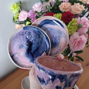 Może przedstawiać: Zestaw ręcznie robionych ceramicznych misek i talerzy z unikalnym marmurkowym wzorem w odcieniach niebieskiego, różowego i fioletowego. Ceramika ma nakrapianą fakturę. Prezentowane na tle kolorowej kompozycji kwiatowej.