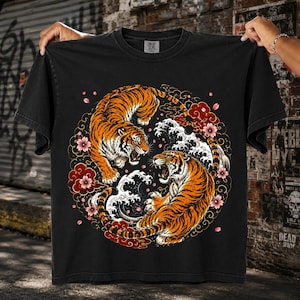 Puede incluir: Una camiseta negra de manga corta con un gráfico circular de dos tigres naranjas y negros frente a frente. El diseño incluye olas blancas estilizadas, nubes rojas y flores de cerezo rosas dispuestas en un patrón circular en el pecho.