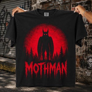 May include: A black short-sleeve t-shirt featuring a graphic of a winged creature standing in front of a large red moon and a forest silhouette. The word MOTHMAN is printed in a distressed red font at the bottom of the graphic design.