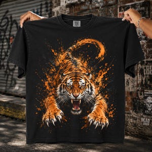 Puede incluir: Una camiseta negra de manga corta con un gráfico grande y detallado de un tigre gruñendo en posición de salto. El tigre está representado en tonos naranja y negro con un efecto dinámico de salpicaduras de pintura alrededor del cuerpo.