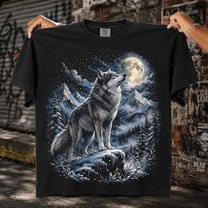 May include: A black short-sleeved t-shirt featuring a graphic print of a wolf standing on a snowy rock. The scene depicts a moonlit night with snow-capped mountains, pine trees, and a starry sky. The shirt has a crew neckline and a Comfort Colors label.