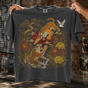 Puede incluir: Una camiseta gris carbón de manga corta con un estampado gráfico de dos tigres naranjas en una pose dinámica. El diseño incluye un sol rojo, flores amarillas y rojas, tallos de bambú y una grulla blanca sobre un fondo oscuro.
