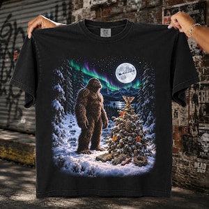 May include: A black short-sleeve t-shirt featuring a graphic print of a brown Bigfoot standing in a snowy forest at night. The scene includes a decorated Christmas tree, a full moon with a silhouette of Santa's sleigh, and green aurora borealis.