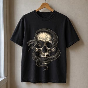 Könnte beinhalten: Ein schwarzes T-Shirt mit kurzen Ärmeln und einem detaillierten Grafikdruck auf der Vorderseite. Das Motiv zeigt einen beigen menschlichen Schädel, der von einer dunklen, gewundenen Schlange umschlungen ist. Der Schlangenkopf ist nahe der Augenhöhle.