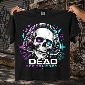 May include: A black t-shirt featuring a graphic of a skull wearing headphones with glowing purple and blue eyes. The design includes musical notes, lightning bolts, and speakers, with the text "DEAD FREQUENCY" printed in white below the skull.