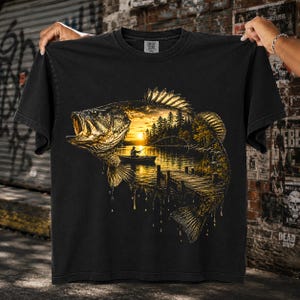 May include: A black short-sleeve t-shirt featuring a large graphic of a largemouth bass fish. Inside the silhouette of the fish is a golden-hued landscape scene of a person in a small boat fishing on a lake at sunset, with pine trees and a dock.