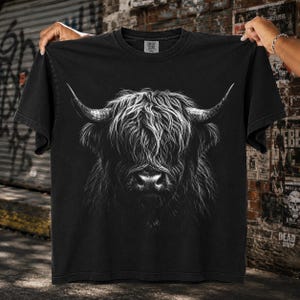 May include: A black short-sleeve t-shirt featuring a detailed white graphic print of a Highland cow head with long, shaggy fur and prominent curved horns. The design is centered on the chest, showcasing the animal's face with a dark, moody aesthetic.
