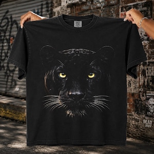 Könnte beinhalten: Ein schwarzes Kurzarm-T-Shirt mit einer großen, detaillierten Grafik eines schwarzen Panthergesichts mit stechend gelben Augen, die mittig auf der Brust platziert ist. Das Shirt hat einen Rundhalsausschnitt und eine verwaschene Vintage-Optik.