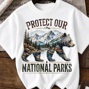 May include: White t-shirt featuring a bear silhouette with a landscape design. The design includes mountains, trees, and the text "PROTECT OUR NATIONAL PARKS" in a distressed font. The shirt is on a hanger.