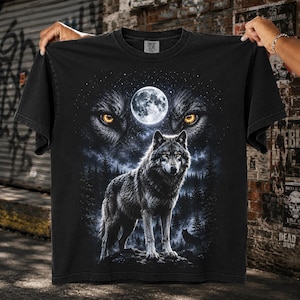 May include: A black t-shirt featuring a graphic print of a wolf standing in a forest at night under a full moon. The design includes a large, detailed wolf face with glowing yellow eyes in the background, set against a starry night sky.
