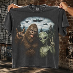 May include: A charcoal gray short-sleeve t-shirt featuring a graphic print of a brown Bigfoot making a rock-and-roll hand gesture and a green alien making a peace sign. Above them, three flying saucers hover over a dark forest landscape.
