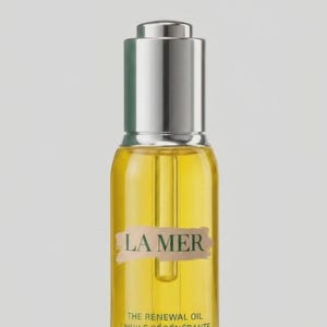 May include: A clear glass bottle filled with golden yellow oil, featuring a silver dropper cap. The label displays the text LA MER, THE RENEWAL OIL, and L'HUILE R&Eacute;G&Eacute;N&Eacute;RANTE in dark green lettering over a light beige brushstroke background.