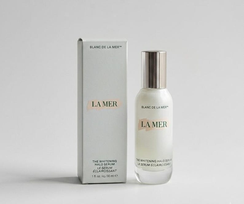 May include: A white rectangular box and a matching glass bottle of La Mer The Whitening Halo Serum. The packaging features the text Blanc de La Mer, La Mer, and The Whitening Halo Serum Le Serum Eclaircissant with a volume of 1 fl. oz./30 ml.