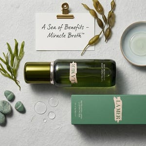 以下が含まれることがあります： La Mer The Treatment Lotionのダークグリーンのガラスボトルと緑色の箱、A Sea of Benefits - Miracle Brothと書かれた小さな白いカード、緑色の海藻の小枝、滑らかな緑色の石、小さな陶器の皿、金色のスプーン。