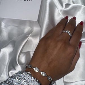 Puede incluir: Una mano con un anillo y una pulsera de diamantes, con una manga plateada con lentejuelas. Una caja blanca con la palabra "GIVENCHY" es visible. El fondo es una tela blanca y sedosa.