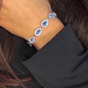 Puede incluir: Una pulsera de plata con piedras preciosas azules claras en forma de pera, rodeadas por un halo de piedras más pequeñas y transparentes. La pulsera se lleva en una muñeca, con una manga de chaqueta negra en el fondo. Joyería.