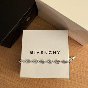 Puede incluir: Una pulsera plateada con piedras preciosas azules ovaladas se exhibe en una caja blanca con el nombre de la marca "GIVENCHY" impreso en negro. Dos cajas de marca están en el fondo. La pulsera está sobre una superficie de madera.