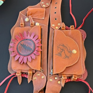 May include: Brown leather chest harness with multiple pockets and straps. One pocket features a red and black sunburst design, while another displays a black graphic. The harness has brass hardware and red accents, with a braided leather strap.
