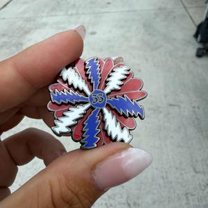 May include: A colorful pin with a floral design. The pin features red, white, and blue segments radiating from a blue center with the number 33. The pin is held between fingers.