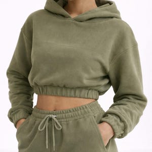 May include: Olive green cropped hoodie and matching sweatpants. The hoodie has a drawstring hood and elasticated cuffs. The sweatpants have a drawstring waist and pockets. The outfit is made of a soft, comfortable material.