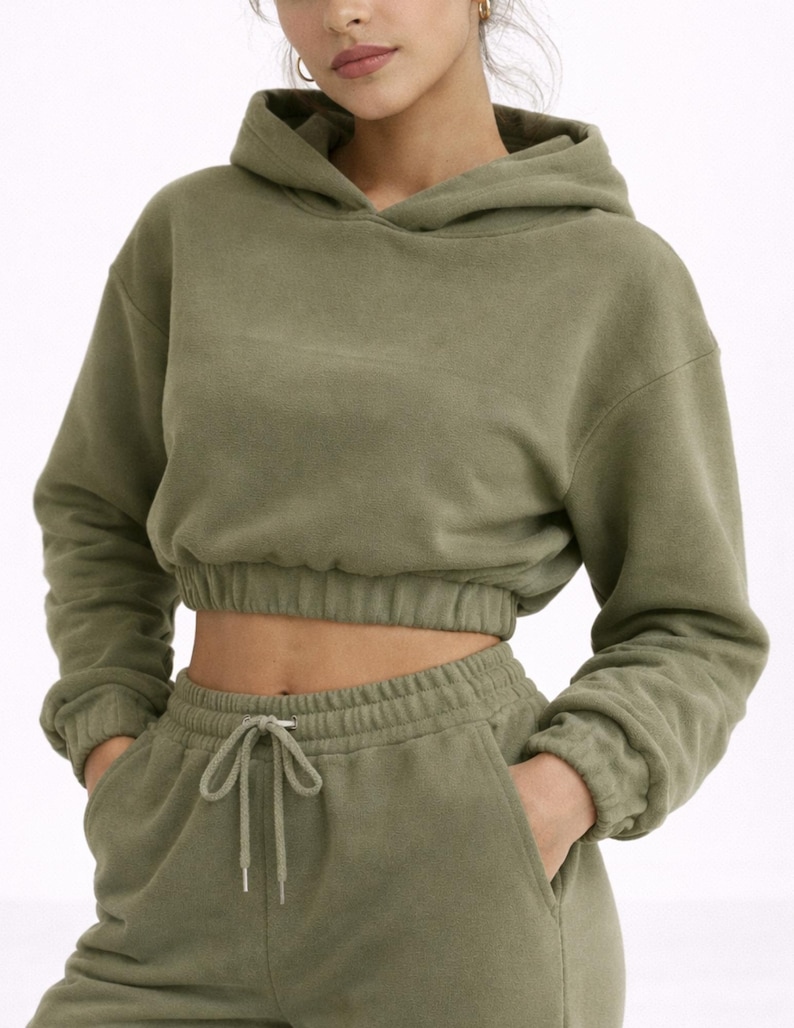May include: Olive green cropped hoodie and matching sweatpants. The hoodie has a drawstring hood and elasticated cuffs. The sweatpants have a drawstring waist and pockets. The outfit is made of a soft, comfortable material.