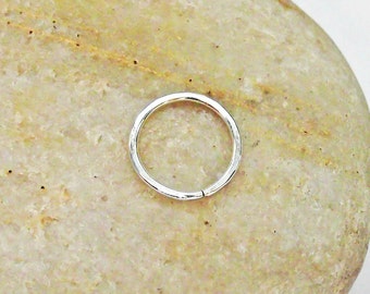May include: A small, silver-colored hoop earring with a simple, minimalist design. The earring is a complete circle with a small gap for insertion. The earring is shown against a light brown, textured background.