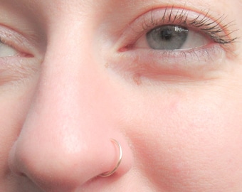 May include: Close-up of a person's face with a small gold nose ring. The person has light skin and blue eyes.