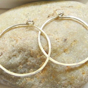 May include: Two silver hoop earrings with a hammered texture. The earrings are on a light brown rock.