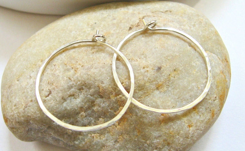 May include: Two silver hoop earrings with a hammered texture. The earrings are on a light brown rock.
