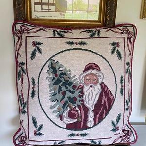 May include: A square tapestry throw pillow featuring a central circular illustration of Santa Claus holding a decorated Christmas tree. The cream-colored fabric is bordered by a dark red trim and decorated with green holly leaves and red bows.